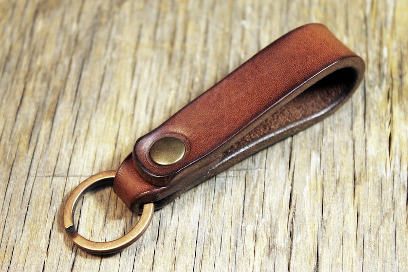 May include: A brown leather keychain with a brass-colored key ring and a brass button closure. The leather is a rich, warm brown color, and the key ring is attached to a loop of the same leather. The keychain is shown on a wooden surface.