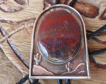 May include: A silver bolo tie with a red, brown, and white agate stone set in a silver frame. The frame features a crescent moon and two cacti. The bolo tie is on a black leather cord.