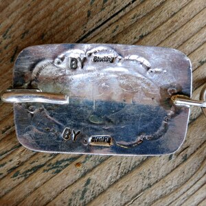 May include: A silver-toned pendant with a rectangular shape and a smooth, polished surface. The pendant has a loop at the top for attaching to a chain. The back of the pendant is stamped with the words "BY Sterling" twice.