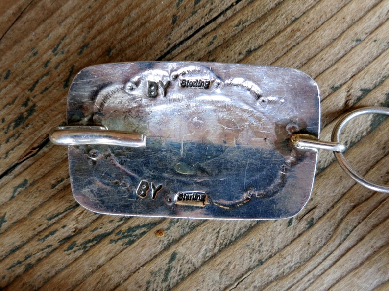 May include: A silver-toned pendant with a rectangular shape and a smooth, polished surface. The pendant has a loop at the top for attaching to a chain. The back of the pendant is stamped with the words "BY Sterling" twice.