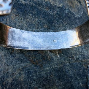 May include: Close-up of a silver bracelet with a detailed design. The inside of the bracelet is stamped with the number "925".