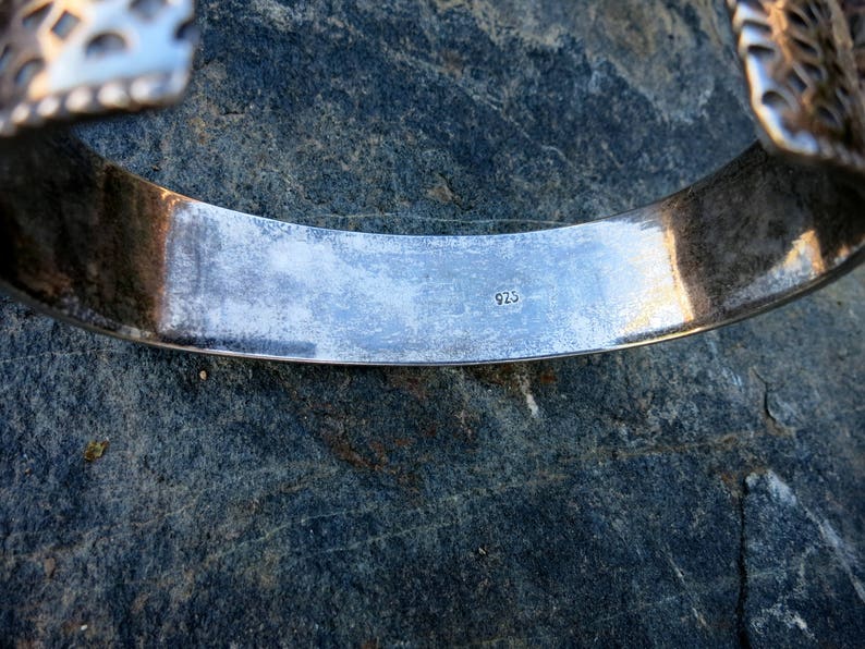 May include: Close-up of a silver bracelet with a detailed design. The inside of the bracelet is stamped with the number "925".