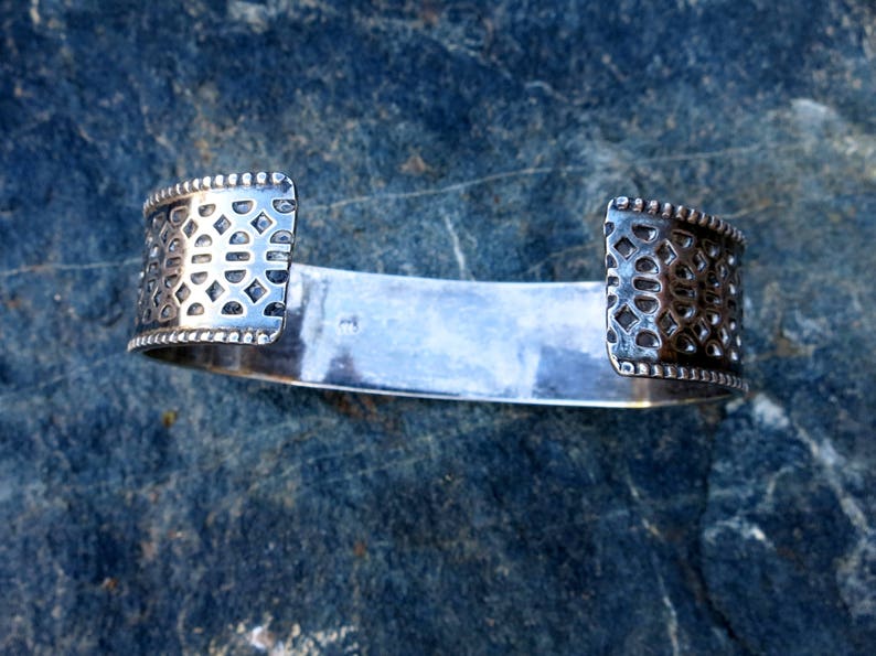 May include: A silver bracelet with a geometric pattern on the ends. The bracelet is open and has a smooth, polished surface.