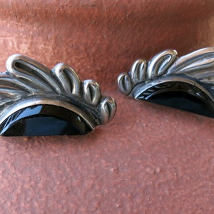 May include: A pair of silver-toned earrings with a stylized leaf design and a black onyx stone. The earrings have a curved shape and are set against a reddish-brown background.