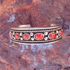 May include: A silver bracelet with a row of red coral stones set in a silver band. The band is decorated with a rope-like design.