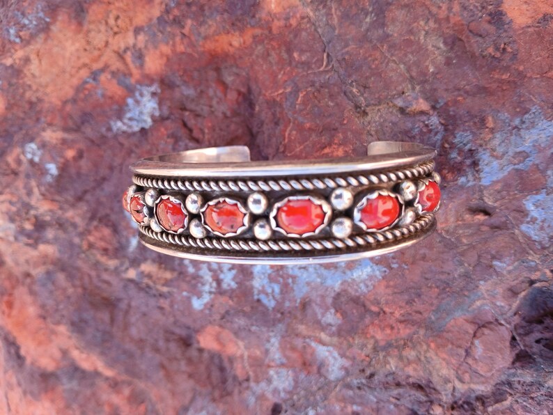 May include: A silver bracelet with a row of red coral stones set in a silver band. The band is decorated with a rope-like design.