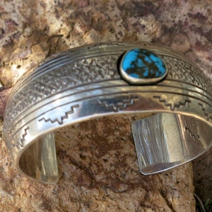 May include: A silver bracelet with a turquoise stone set in the center. The bracelet is decorated with a geometric pattern.