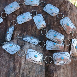 May include: A silver chain belt with turquoise stones set in rectangular silver plaques. The belt is laid out on a rough, brown rock surface.