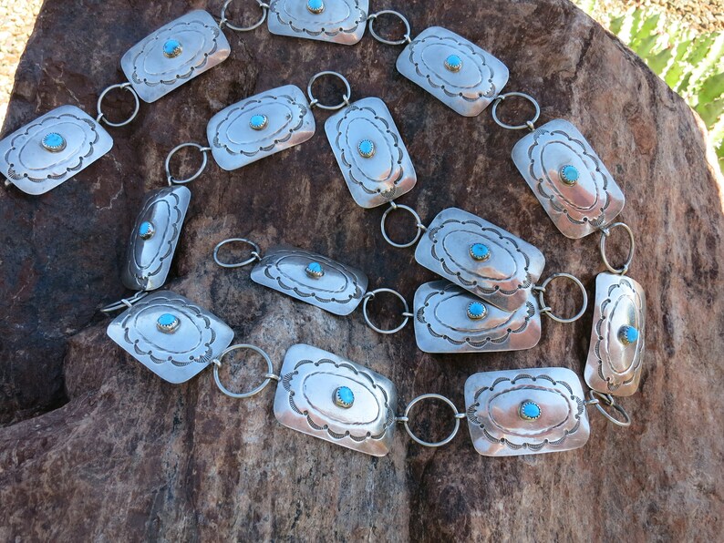May include: A silver chain belt with turquoise stones set in rectangular silver plaques. The belt is laid out on a rough, brown rock surface.