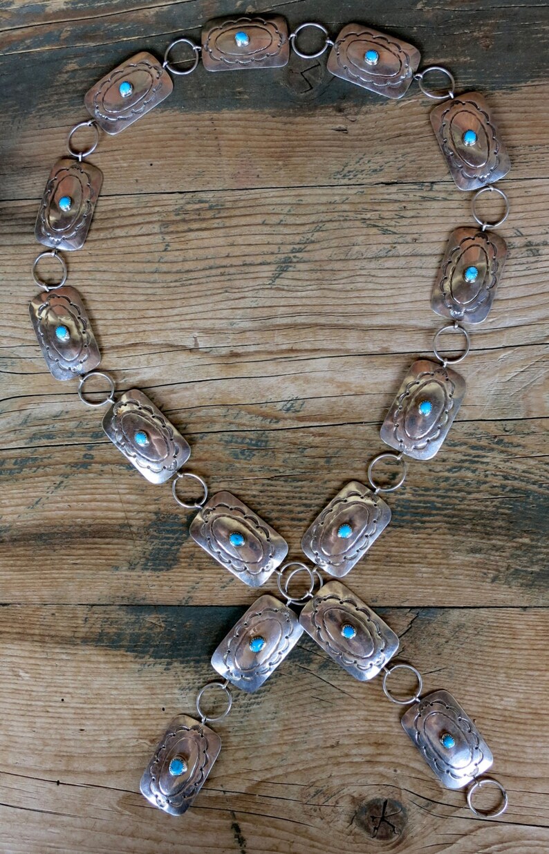 May include: A silver chain belt with rectangular turquoise stones set in each link. The belt is laid out on a wooden surface.