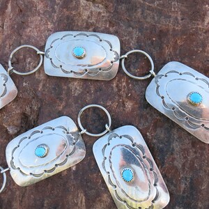 May include: A silver belt with turquoise stones and decorative designs. The belt is made up of rectangular links connected by silver rings.