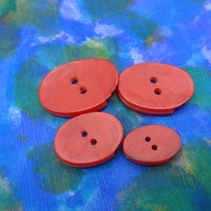 Puede incluir: Cuatro botones rojos ovalados con dos agujeros cada uno, dispuestos sobre una tela estampada azul y verde. Los botones var&iacute;an en tama&ntilde;o, con dos m&aacute;s grandes y dos m&aacute;s peque&ntilde;os. Los botones tienen un borde ligeramente elevado.