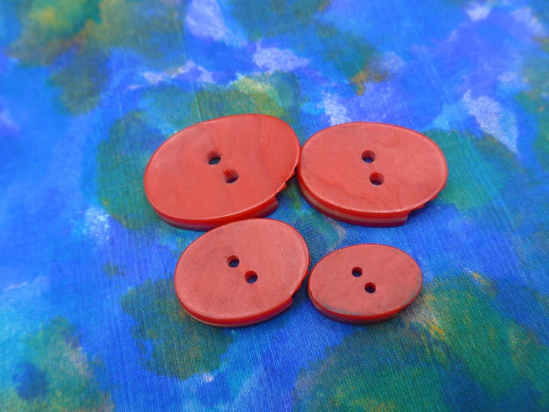 Puede incluir: Cuatro botones rojos ovalados con dos agujeros cada uno, dispuestos sobre una tela estampada azul y verde. Los botones var&iacute;an en tama&ntilde;o, con dos m&aacute;s grandes y dos m&aacute;s peque&ntilde;os. Los botones tienen un borde ligeramente elevado.