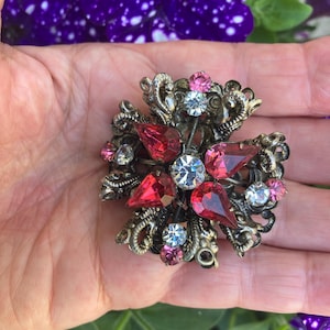 May include: Vintage brooch with a cross-like design. The brooch features a bronze-colored metal base adorned with red, pink, and clear rhinestones. The central stone is a large, clear, round rhinestone, surrounded by smaller stones.
