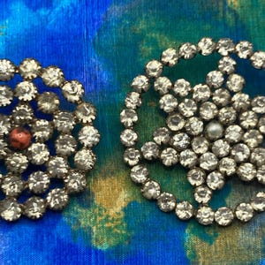May include: Two vintage rhinestone brooches. One is a circular design with a red center stone, and the other is a star shape with a pearl center. Both are set against a blue and green patterned background.