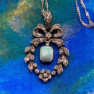 May include: Silver-toned pendant necklace featuring a bow, a wreath of leaves, and a floral design. The pendant has a rectangular, light blue stone at its center, and is encrusted with small, clear gemstones. The necklace chain is also silver-toned.