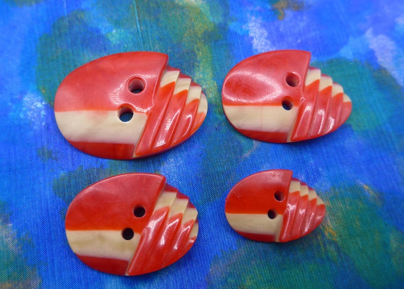 Puede incluir: Cuatro botones ovalados de diferentes tama&ntilde;os. Cada bot&oacute;n es rojo con una franja crema y un dise&ntilde;o tallado en forma de abanico. Los botones tienen dos agujeros para coser y est&aacute;n sobre un fondo azul.