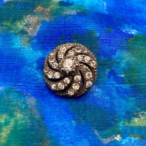 May include: A silver-toned button with a spiral design, encrusted with small, clear gemstones. A larger, clear gemstone is set in the center. The button is set against a blue and green patterned fabric.