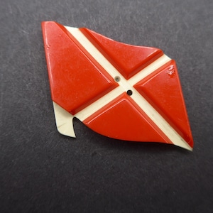 May include: A vintage red and cream-colored flag-shaped button. The button features a cross design with a small hole in the center for sewing. The button has a glossy finish and is set against a dark background.