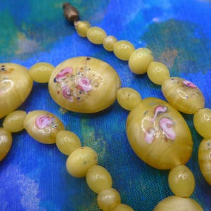 May include: A vintage necklace with yellow oval beads and larger, flattened beads. The flattened beads are decorated with hand-painted pink and white floral designs. The necklace is displayed on a blue and green patterned fabric.