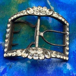 May include: A rectangular metal shoe buckle featuring a curved frame adorned with numerous clear, faceted rhinestones. The buckle includes a central hinged prong mechanism and two smaller prongs on the side, all set within a metallic frame.