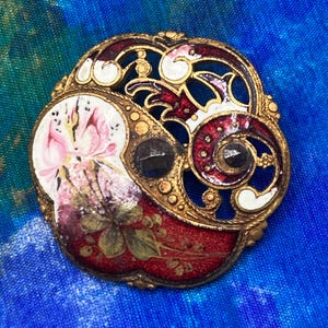 May include: A round gold-tone brooch featuring intricate openwork metal filigree, red and white enamel detailing, and a hand-painted floral motif on a white background. The piece is accented with two dark, faceted circular stones set within the frame.