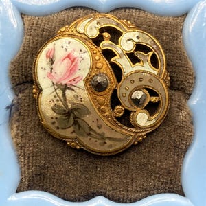 May include: A circular gold-tone brooch featuring a painted pink rose on a cream-colored enamel background on the left side, and an openwork filigree design with two small dark inset stones on the right side, all set within a textured metal frame.