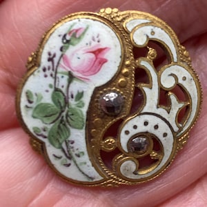 May include: A round gold-tone brooch featuring a white enamel inlay with a hand-painted pink rosebud and green leaves. The metal frame has an intricate openwork scroll design accented with two small, dark, faceted stones set into the surface.