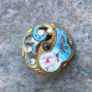 May include: A circular gold-toned metal button featuring intricate openwork scroll designs in light blue enamel. The center displays a small dark stone accent, while one side features a white enamel section painted with a pink floral motif.