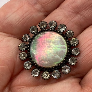 May include: A round brooch featuring a central iridescent cabochon with pink and green hues, surrounded by a halo of small, clear, faceted stones set in a dark metal frame. The circular design resembles a sunburst with a shimmering centre.