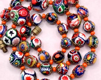May include: A necklace featuring multi-colored millefiori glass beads with intricate floral and star patterns. The round beads are separated by small, bright orange spacer beads and accented with antique-style brass metal bead caps.