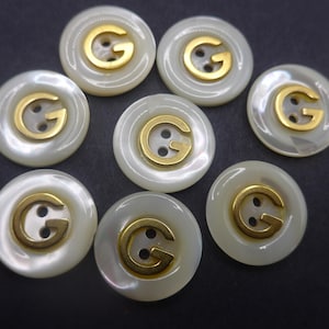 May include: Eight round, off-white buttons with a gold-coloured "G" emblem in the centre. Each button has two holes for sewing. The buttons are arranged on a dark surface.