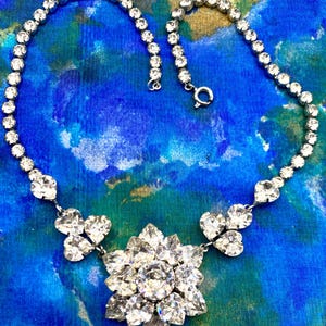 May include: A silver-toned necklace with clear, sparkling rhinestones. The necklace features a central floral pendant and smaller heart-shaped clusters. The chain is composed of small, round rhinestones. The background is a blue and green patterned fabric.