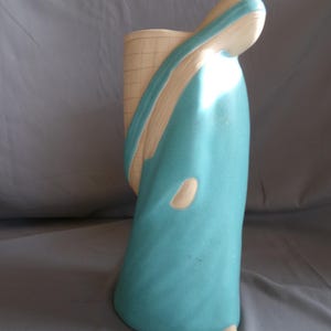 May include: A teal ceramic vase with a curved design. The vase features a light beige accent on the top and a grid pattern on the back. The vase has a smooth, glossy finish and a unique, artistic shape.
