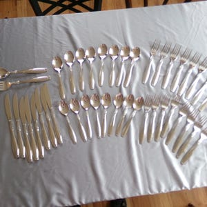 May include: A complete set of silver-colored flatware arranged on a gray tablecloth. The set includes spoons, forks, and knives, ideal for a formal dining setting. The silverware is arranged in a fan shape.