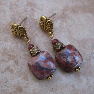 May include: A pair of dangle earrings featuring reddish-brown, square-shaped stones with dark veining. The earrings have gold-colored metal accents, including ornate stud tops, decorative caps, and small chain links. The earrings are designed for pierced ears.
