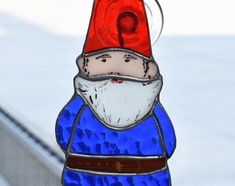 May include: A stained glass gnome with a red pointed hat and a blue tunic. The gnome has a white beard, brown boots, and a brown belt. The glass is held together with dark metal lines. A suction cup is attached to the back.