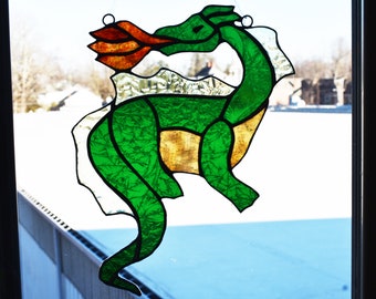May include: A stained glass dragon in shades of green, gold, and orange. The dragon is depicted in a side profile, with its mouth open, exhaling orange flames. The glass is outlined in black, and the background is a snowy outdoor scene.
