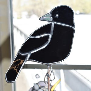 May include: A stained glass bird sculpture featuring a black body with silver-colored outlines. The bird has a light green beak and a copper-colored wire stand. The tail feathers have a brown and gold pattern.
