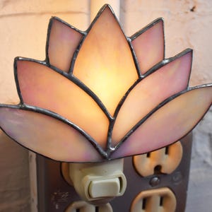 May include: A stained glass night light shaped like a lotus flower with iridescent pale pink petals and dark metal soldering. The light is plugged into a standard wall outlet, emitting a warm glow from behind the translucent glass segments.