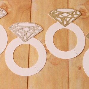 May include: Four white paper ring cutouts with glittery diamond shapes on a wooden surface. The diamonds have a silver or gold glitter finish. The rings are arranged in a row, suggesting a wedding or engagement theme.