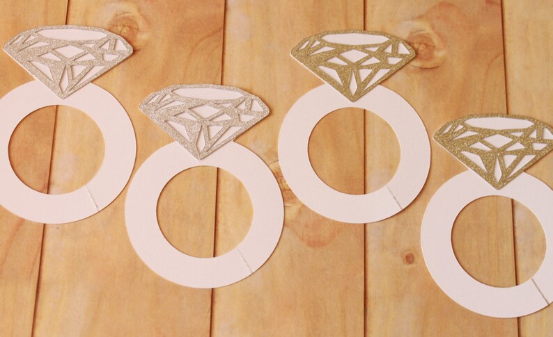May include: Four white paper ring cutouts with glittery diamond shapes on a wooden surface. The diamonds have a silver or gold glitter finish. The rings are arranged in a row, suggesting a wedding or engagement theme.