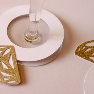 May include: A close-up of a champagne glass with a white base and gold glitter diamond-shaped decorations. The diamonds are cut out with geometric patterns. The background is a soft pink.