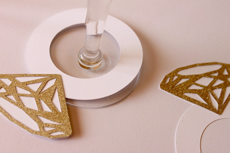 May include: A close-up of a champagne glass with a white base and gold glitter diamond-shaped decorations. The diamonds are cut out with geometric patterns. The background is a soft pink.