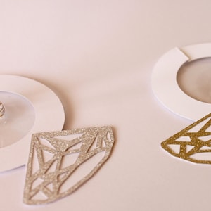 May include: Close-up of two wine glasses with white paper coasters. Two diamond-shaped glittery ornaments, one silver and one gold, are placed near the glasses. The background is a soft, off-white color.