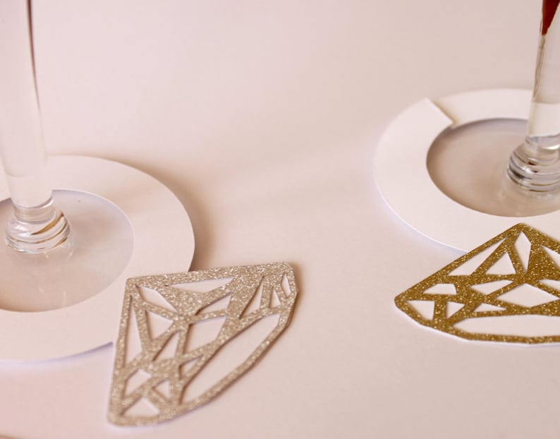 May include: Close-up of two wine glasses with white paper coasters. Two diamond-shaped glittery ornaments, one silver and one gold, are placed near the glasses. The background is a soft, off-white color.