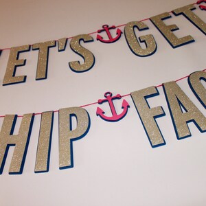 May include: A two-line banner featuring gold glitter letters spelling out LET'S GET SHIP FACED. The letters are mounted on a dark blue backing and are strung together with a thin pink cord. Small pink anchor cutouts are placed between the words.