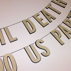 May include: A banner with the words "TIL DEATH DO US PARTY" in glittery gold letters. The letters are strung on a black string against a white background. Ideal for wedding or party decorations.