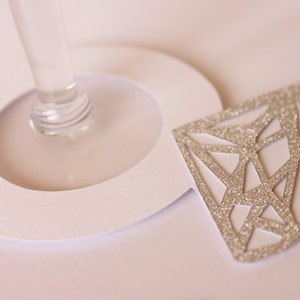 May include: A close-up of a champagne glass with a white paper cutout and a glittery diamond-shaped decoration. The diamond is silver and has a geometric design. The glass stem is clear.