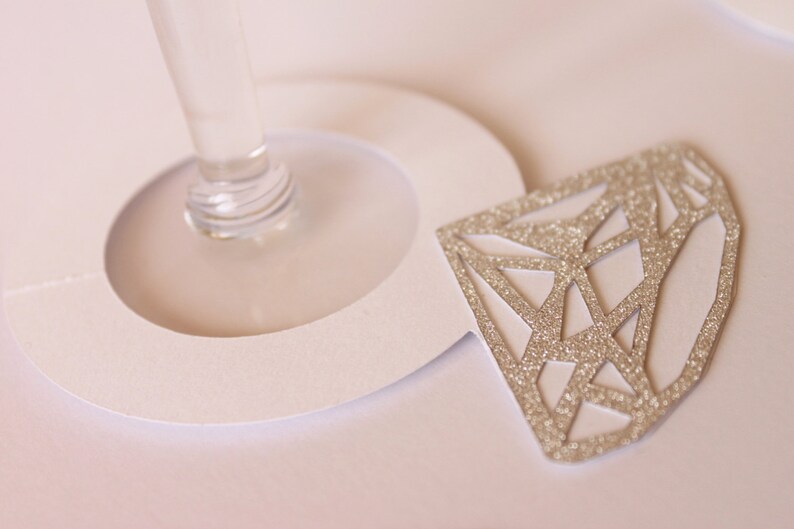 May include: A close-up of a champagne glass with a white paper cutout and a glittery diamond-shaped decoration. The diamond is silver and has a geometric design. The glass stem is clear.